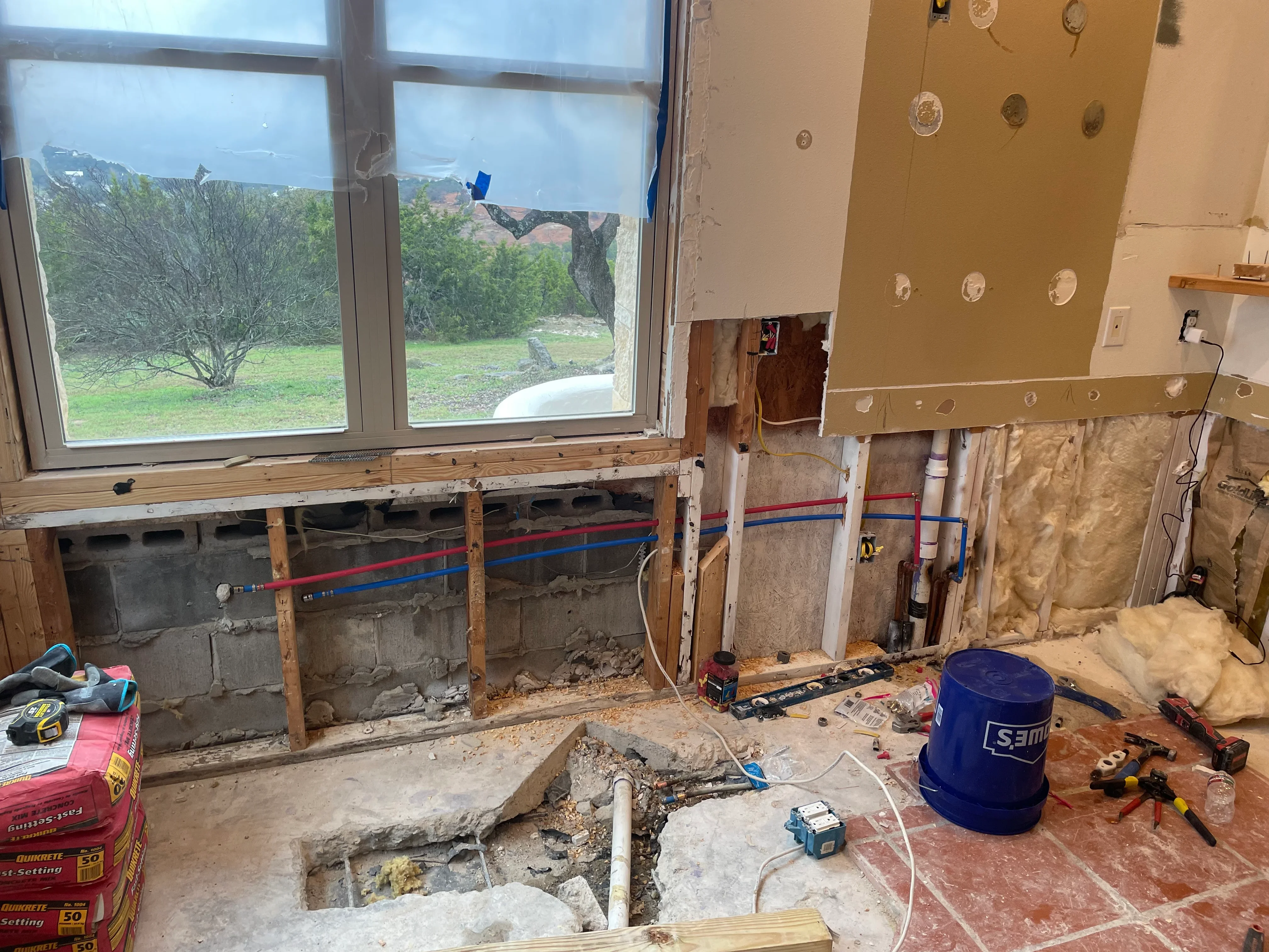 Kitchen renovation in progress showing framed walls, plumbing lines, concrete work, and large windows