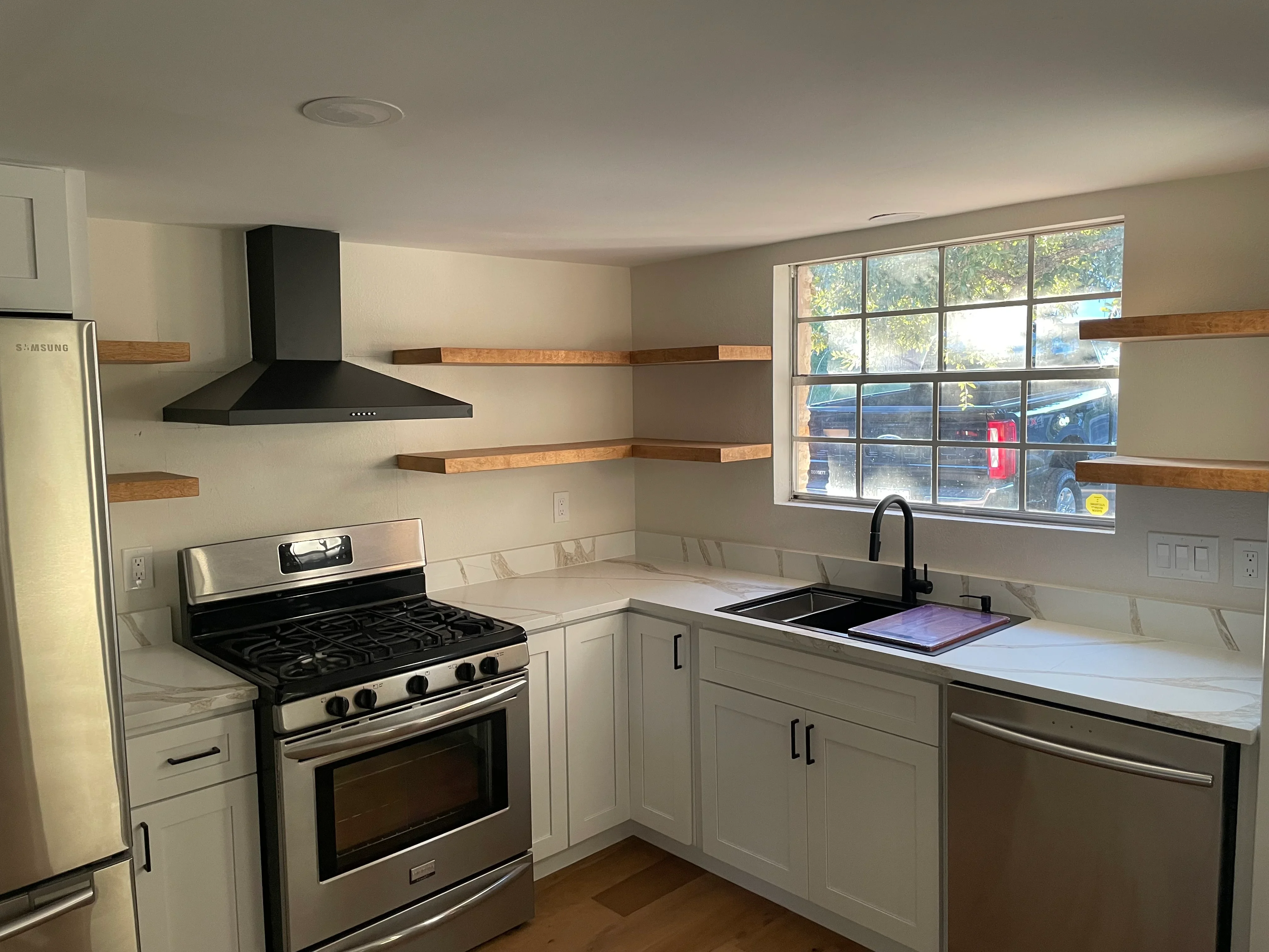 White kitchen cabinets with stainless steel appliances, floating wood shelves, and black range hood