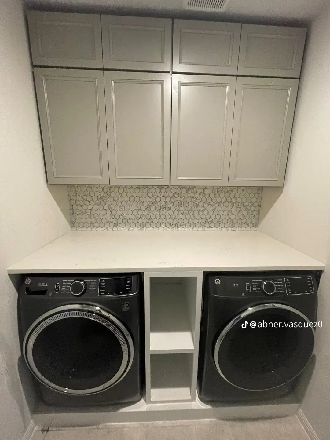 Custom white cabinetry, marble hexagon backsplash, and built-in shelving above dual black front-load washers and dryers