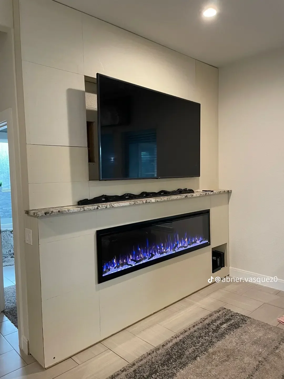 Built-in electric fireplace with blue flames, granite countertop, and wall-mounted TV in cream tile surround