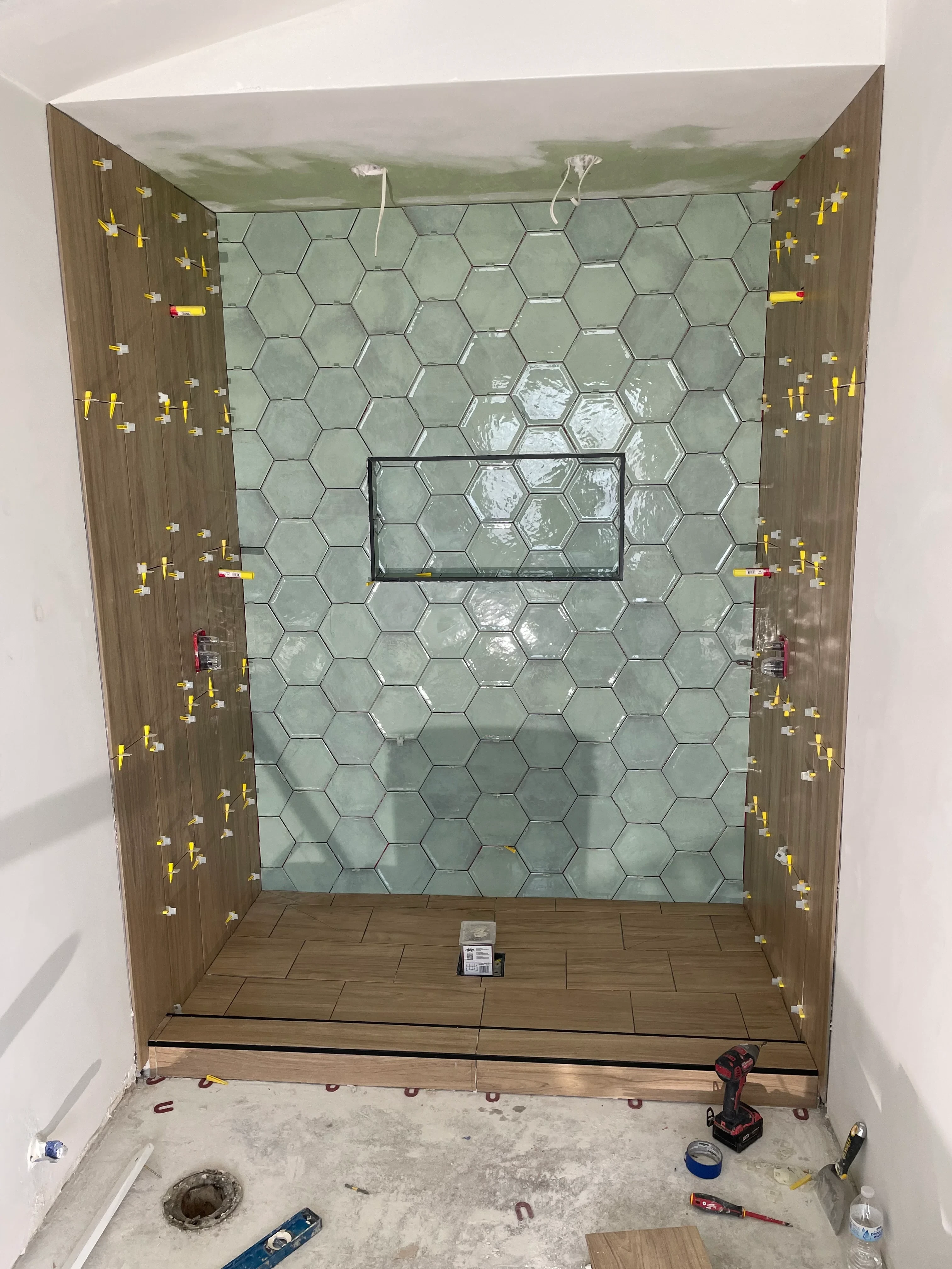 Hexagonal tile shower walls with frameless glass door and wood-look tile flooring during installation