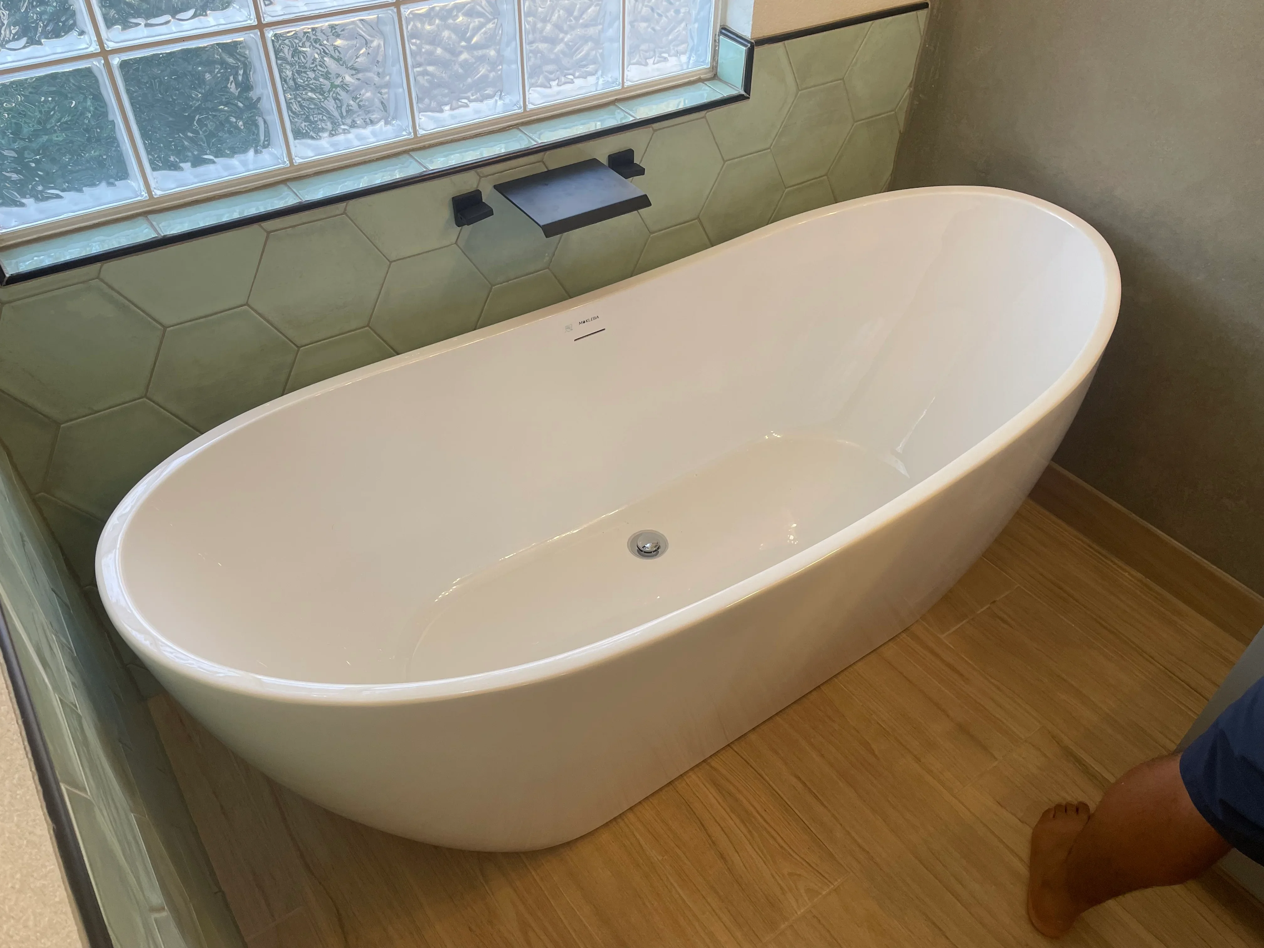 Freestanding white soaking tub with hexagonal tile surround and wood flooring in renovated bathroom